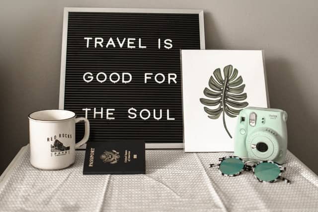 teal fujifilm instax mini camera near white ceramic mug and a sign that says travel is good for the soul